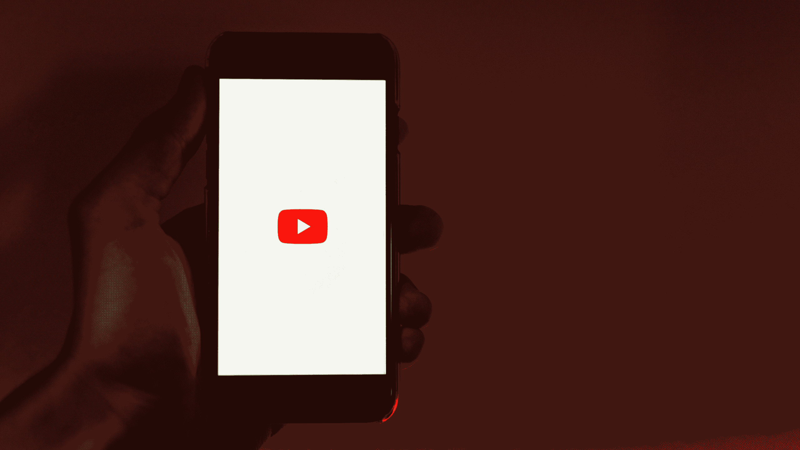 A hand holding a smartphone displaying the YouTube logo on a white screen, with a dark red background.