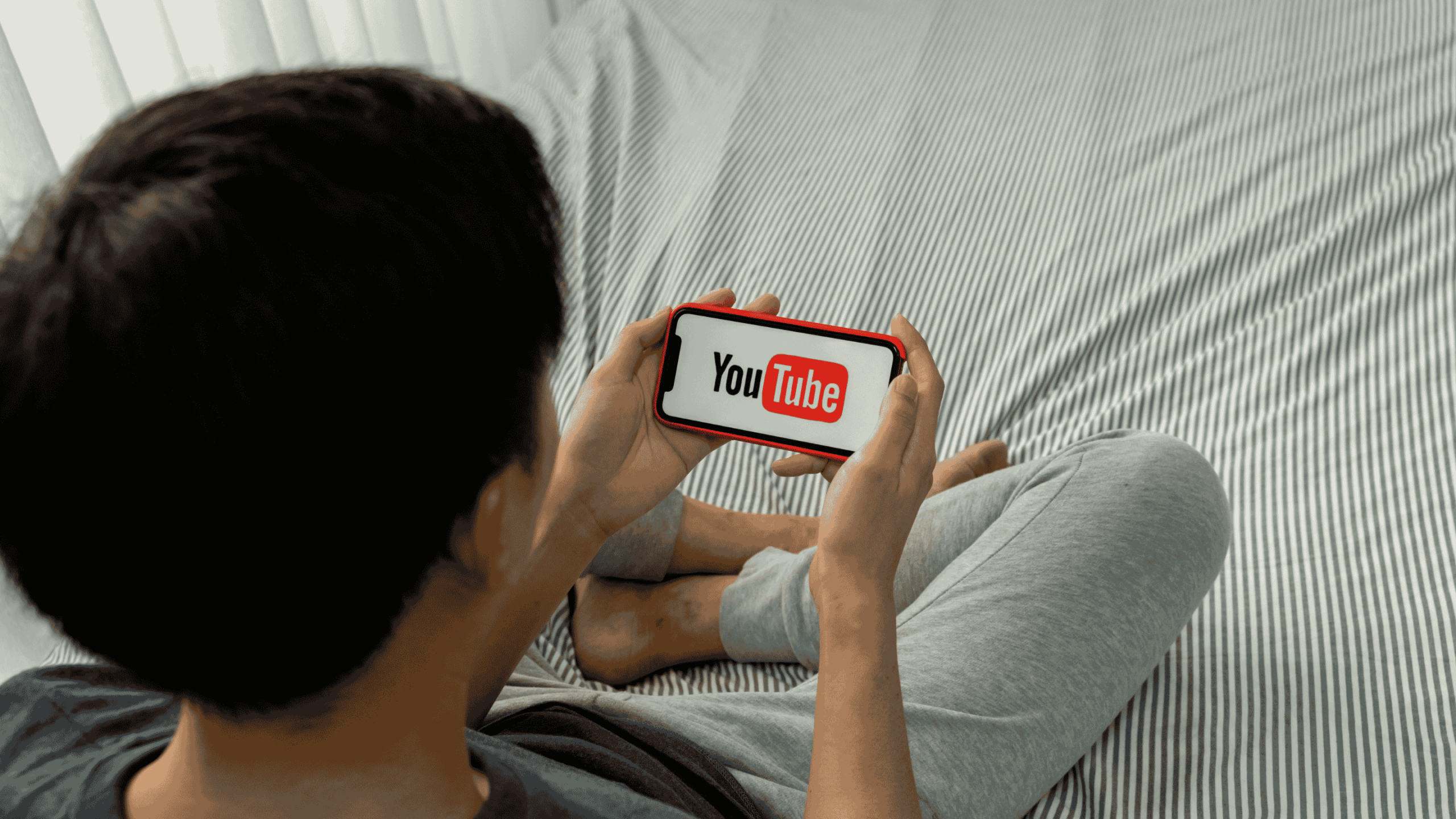 Person sitting on a bed while holding a smartphone showing the YouTube logo on the screen.