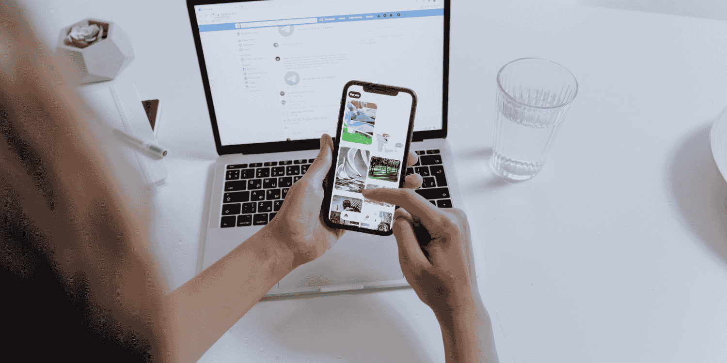 Person browsing Facebook on a smartphone while using a laptop, searching for giveaways through social media posts and Facebook groups.