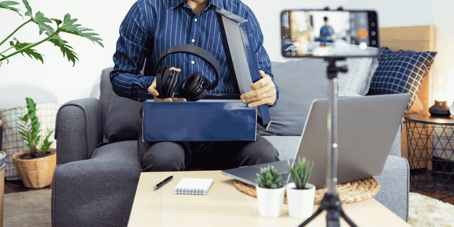 Man unboxing a pair of headphones while live streaming on a smartphone, with a laptop and notepad on the table—representing the topic "How to Find Gleam Giveaways".