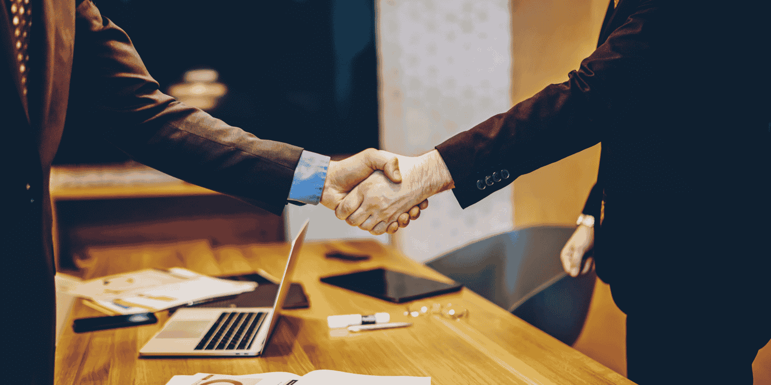 Two professionals in business attire shaking hands across a table with laptops and documents, symbolizing a successful sponsorship deal for a giveaway campaign.
