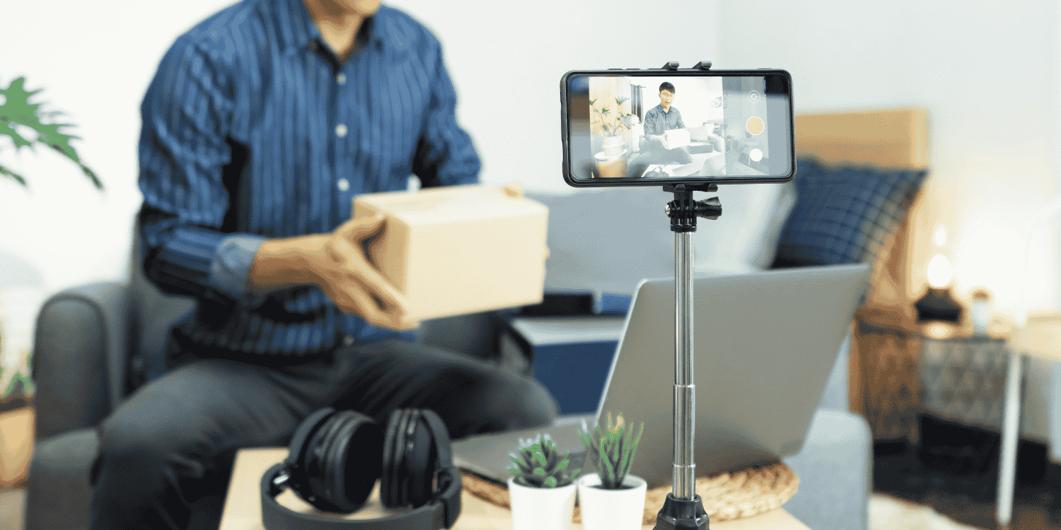Man live streaming from home while holding a giveaway box in front of a smartphone camera, with a laptop and gaming headset nearby—representing the topic "How to Win Giveaways on Twitch"